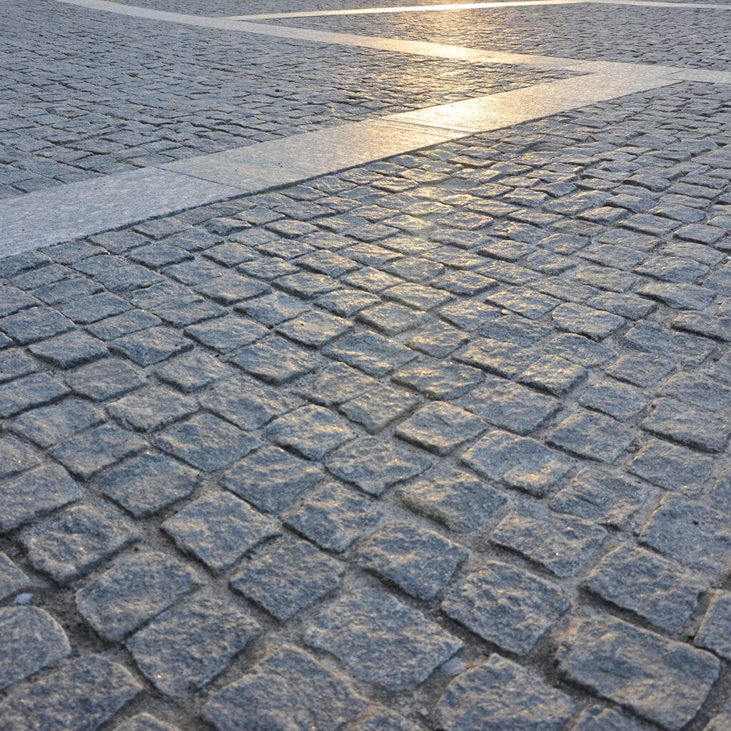 A person walks along a granite cobblestone street as the sun sets in the background, casting a warm glow.