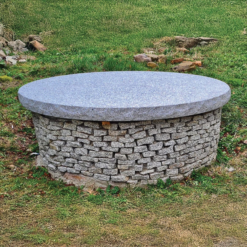 A stone fire pit made of granite cobbles with big granite slab used as cover, elegantly placed in a grassy area, serving as a garden ornament